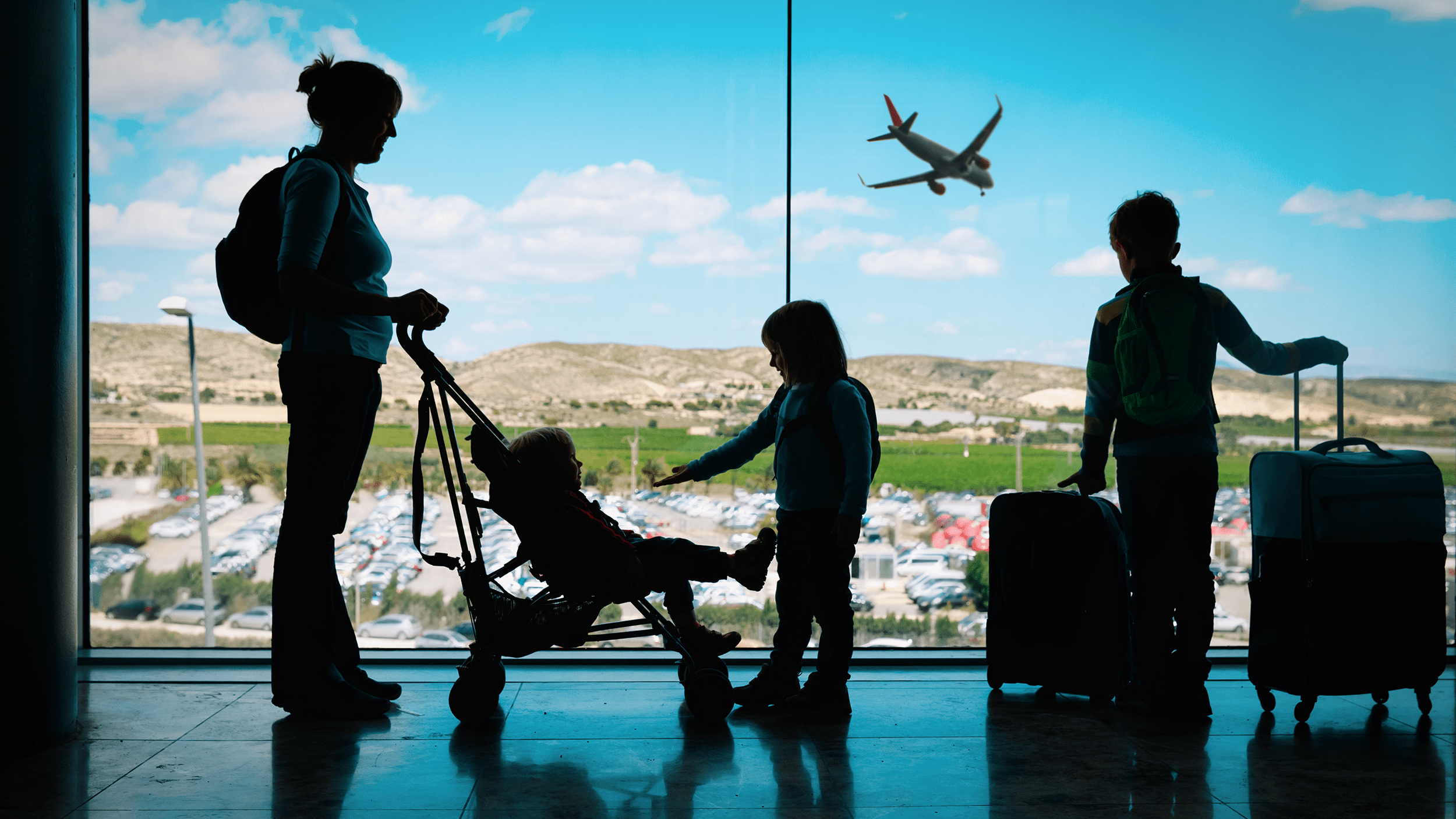 Family at an airport terminal in silhouette, with a stroller and luggage, plane visible outside the window.