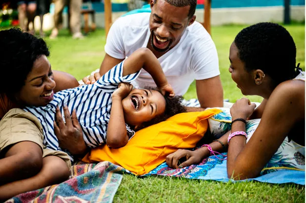 A diverse family plays on a blanket in a park, laughing and enjoying time together with a child in the center.