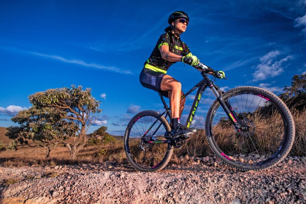 Female mountain biker riding a black and neon-green bike on a rocky desert trail under a blue sky, wearing helmet and sunglasses.