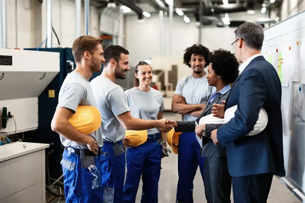 Factory workers in gray shirts and blue coveralls shake hands with a suited supervisor near a whiteboard in a manufacturing plant.