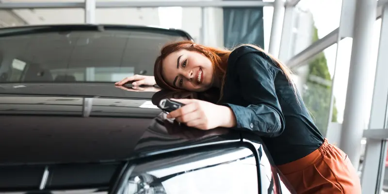 Smiling woman leaning on a black car in a showroom while holding car keys.