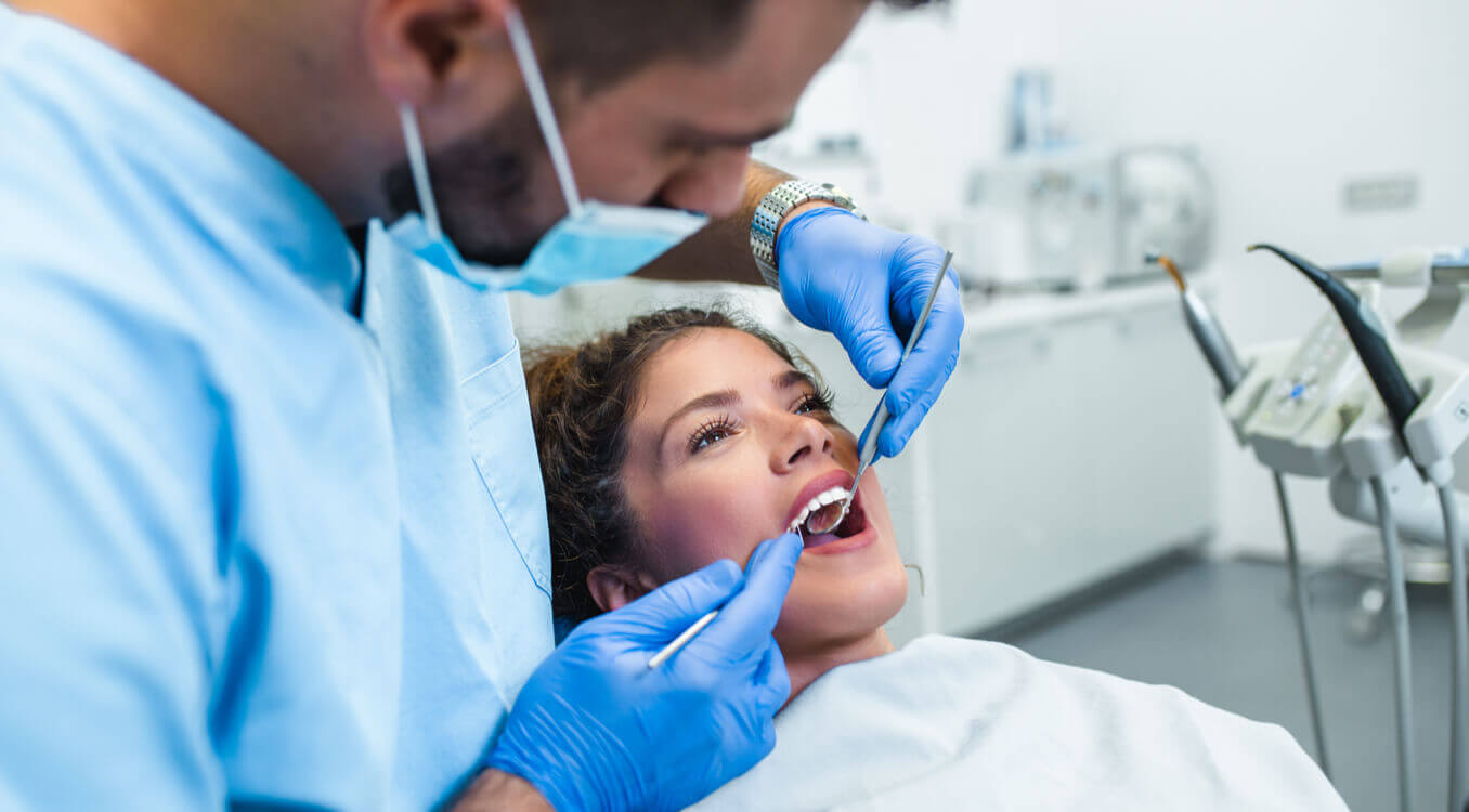 Dentist in blue scrubs and gloves examining a patient’s mouth with dental instruments in a clinic setting.