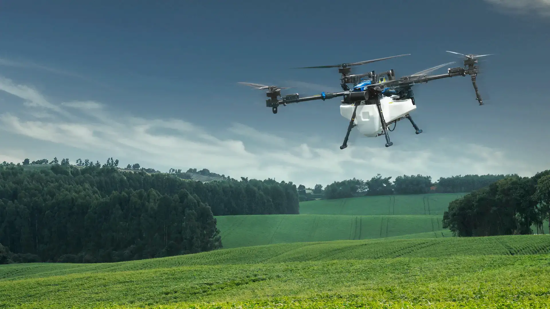 Agricultural drone with a white tank flies over green fields to spray crops.