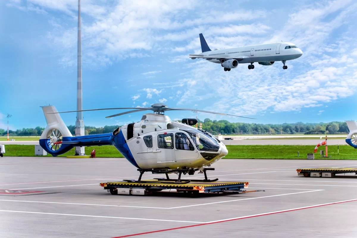 White helicopter parked on a wheeled platform on an airport tarmac with a passenger jet flying overhead against a blue sky.