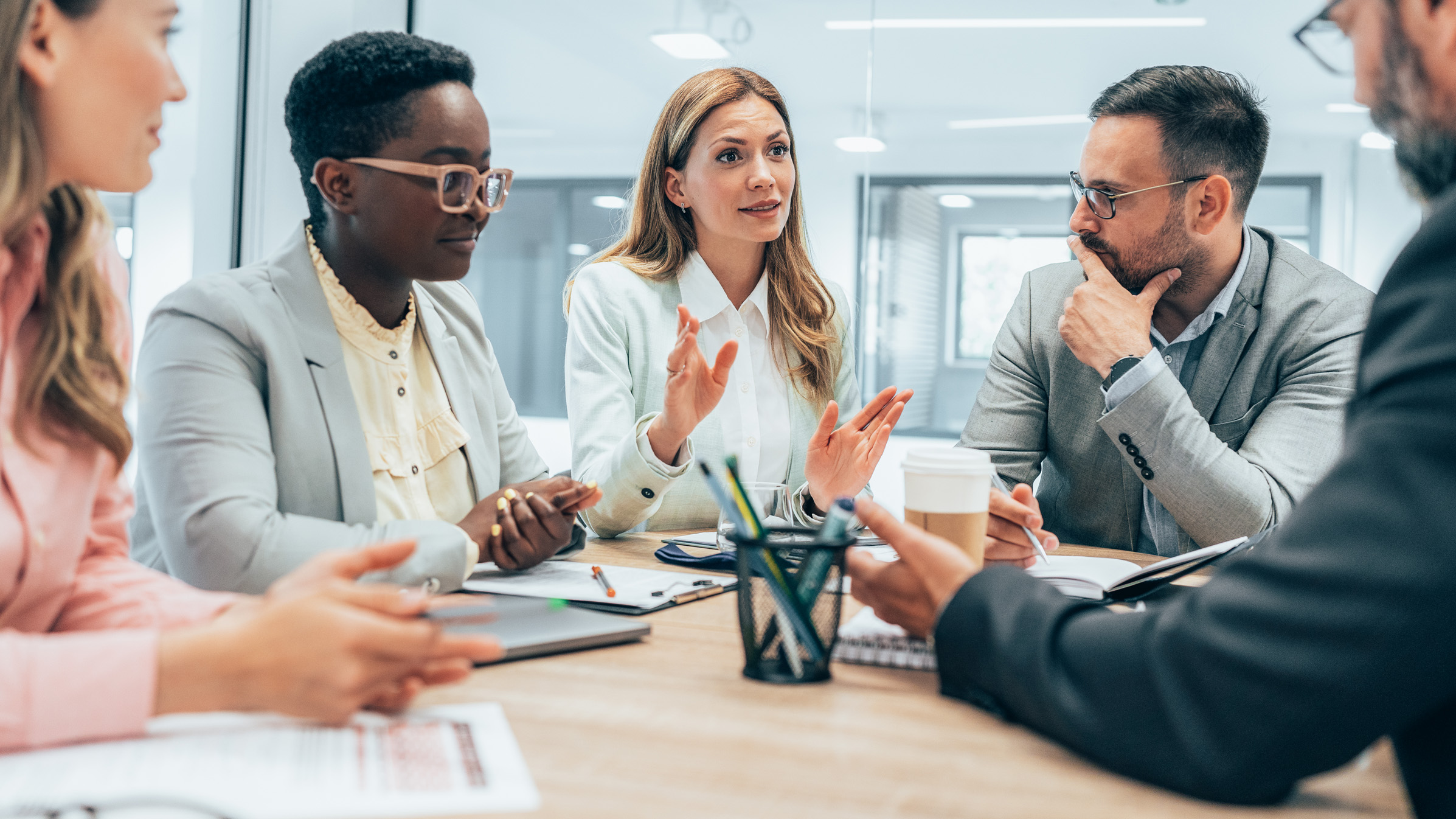 Group of diverse professionals in a business meeting around a table, one woman speaking as others listen and take notes.
