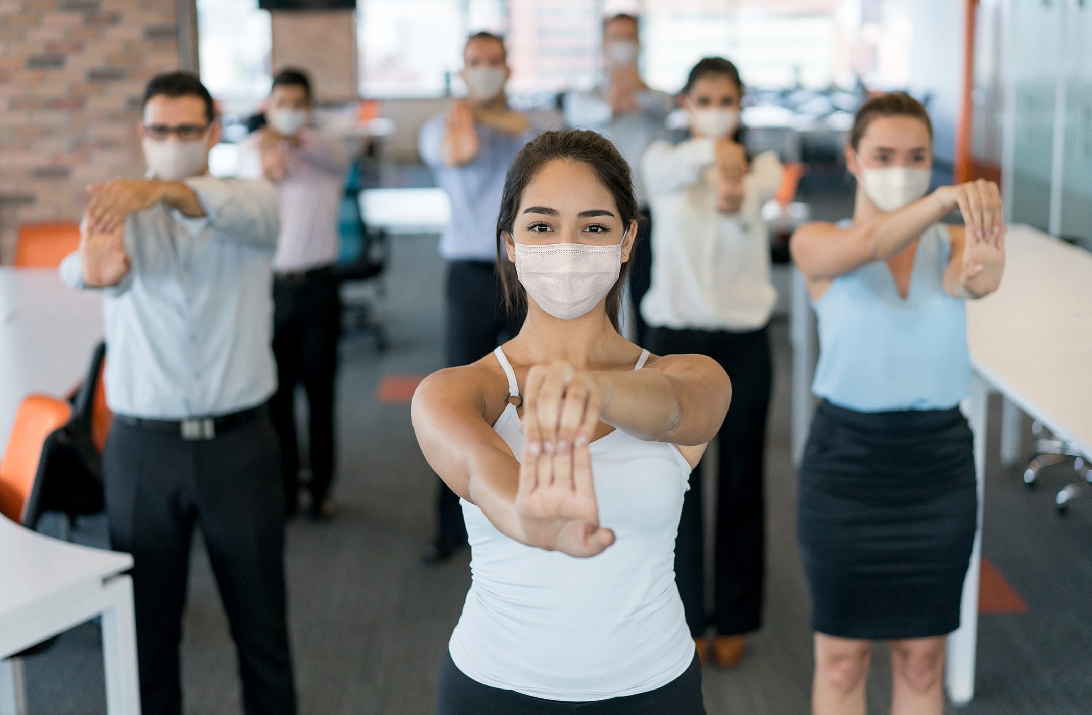 Front-facing woman in a white mask extends her arms forward while coworkers in masks perform similar stretches in a bright office.