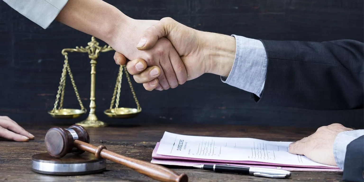 Two people shake hands over a desk with a gavel, documents, and scales of justice in the background.
